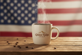 A warm patriotic coffee scene with a steaming mug of coffee on a rustic wooden table, softly blurred American flag in the background. Add a simple mountain-style logo labeled “Grace Ridge Coffee Co.” on the mug in clean brown ink. Premium lifestyle aesthetic, soft natural light, no clutter.